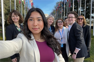 [ai] A group of six women posing for a selfie outdoors, with multiple international flags in the background. The woman in front is wearing a light jacket and looks directly at the camera, while the others stand behind her smiling.
